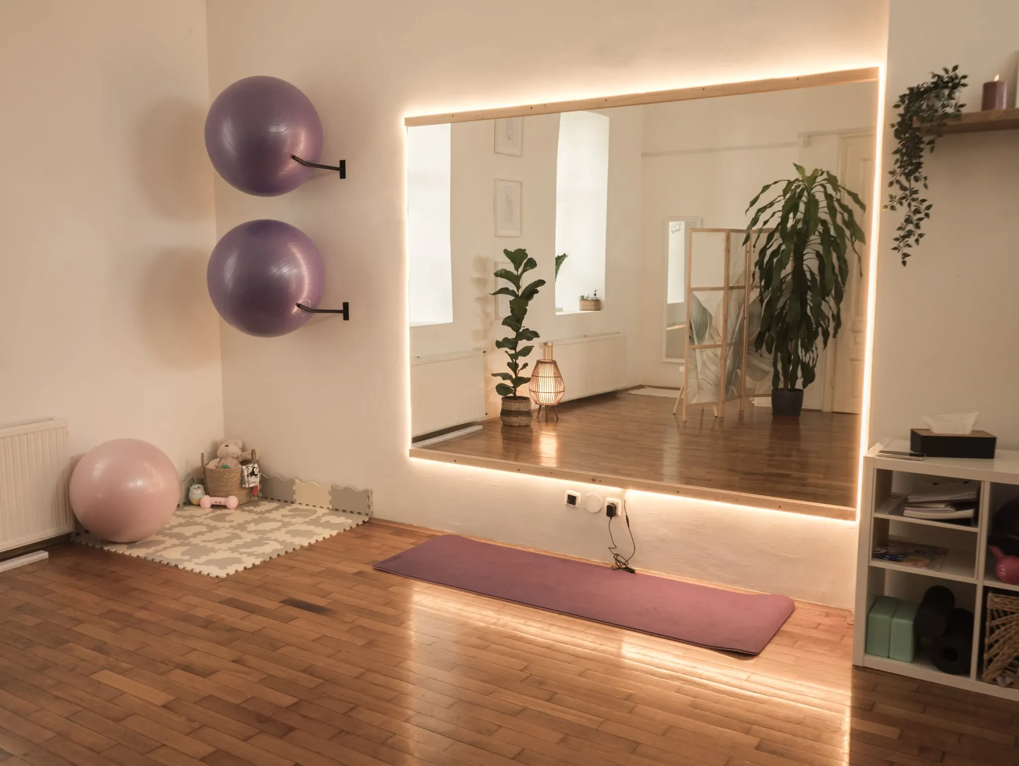A stúdió edzőterme: nagy, hátulról megvilágított tükör, falra akasztott lila fitneszlabdák, rózsaszín nagylabda és edzőmatrac a parkettán